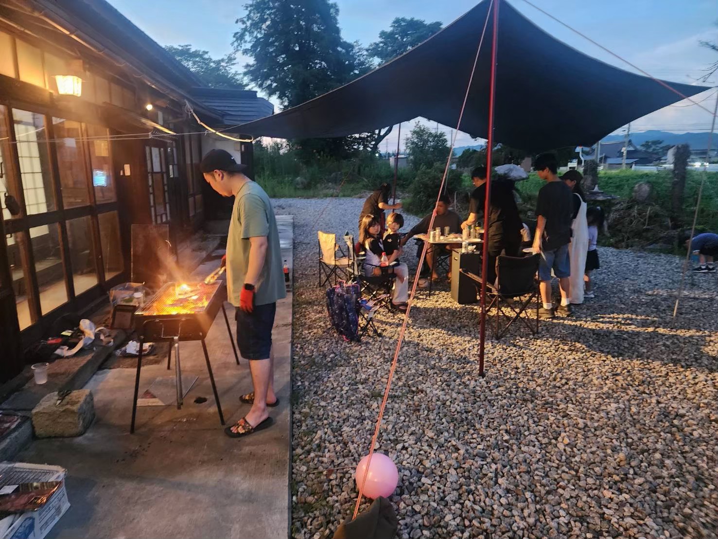 古民家の前でBBQを焼く参加者と、テーブルを囲む大人と子どもたち